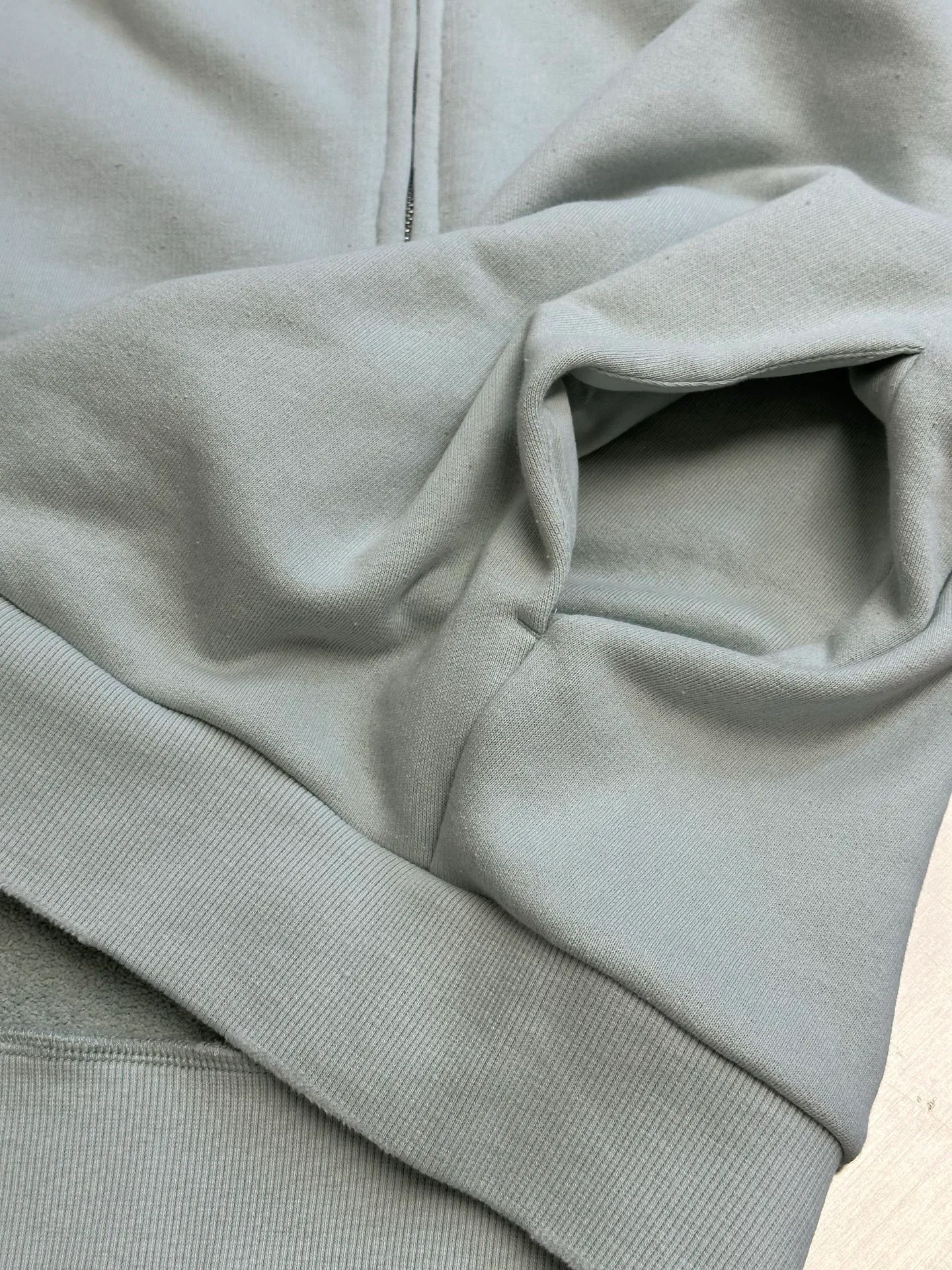 Ac vintage collared zipper sweatshirt uses 32-count top layer and 40-count bottom layer of 3-strand composite fleece structure, with a weight of g/㎡. The 32-count yarn on the top layer is fine and smooth, giving the surface a smooth touch and excellent anti-pilling properties; the 40-count 3-ply yarn on the bottom layer is treated with a raising process to form a rich and soft fluff layer, providing excellent warmth and skin-friendly wrapping. With 21 2×2 ribs, the g/㎡ weight ensures that the collar, cuffs and hem have excellent tensile fatigue resistance and shape retention capabilities. During piece dyeing, the entire fabric is immersed in the dye vat after weaving, and the dye molecules fully penetrate into the fibers, ensuring full, deep colors and excellent color fastness. Each batch of dyeing produces subtle differences, making the color tone of the single product unique. On this basis, the paint is sprayed irregularly on the surface of the garment through manual spray painting to create a splash-like retro effect. The distressed processing of holes causes the color to fade and mottle in parts. The ribbed trim is also treated to form slight whitening and fluffing, which visually echoes the spray-painted effect on the body, making the new clothes appear as if they have been worn for a long time and exposed to the sun. The front logo stamp uses a slurry printing process. The printing slurry contains a special microcapsule foaming agent. After being baked at high temperature, the slurry layer expands to form three-dimensional bulges, and the surface presents a granular texture similar to inkjet paint. This process simulates the effect of random drops of pigment after being atomized during spray gun spraying. The edges of the pattern present a natural feathering transition, rather than the rigid boundaries of glue printing. The printing surface has a fine graininess and soft elasticity to the touch, forming a closed loop of the process with the distressed effect of the spray painting on the body. size：xs s m l